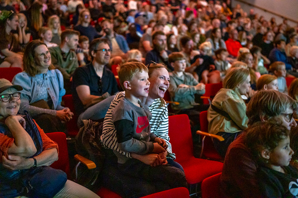 Publikum bl.a. en mor med et barn på skødet griner i en teatersal.